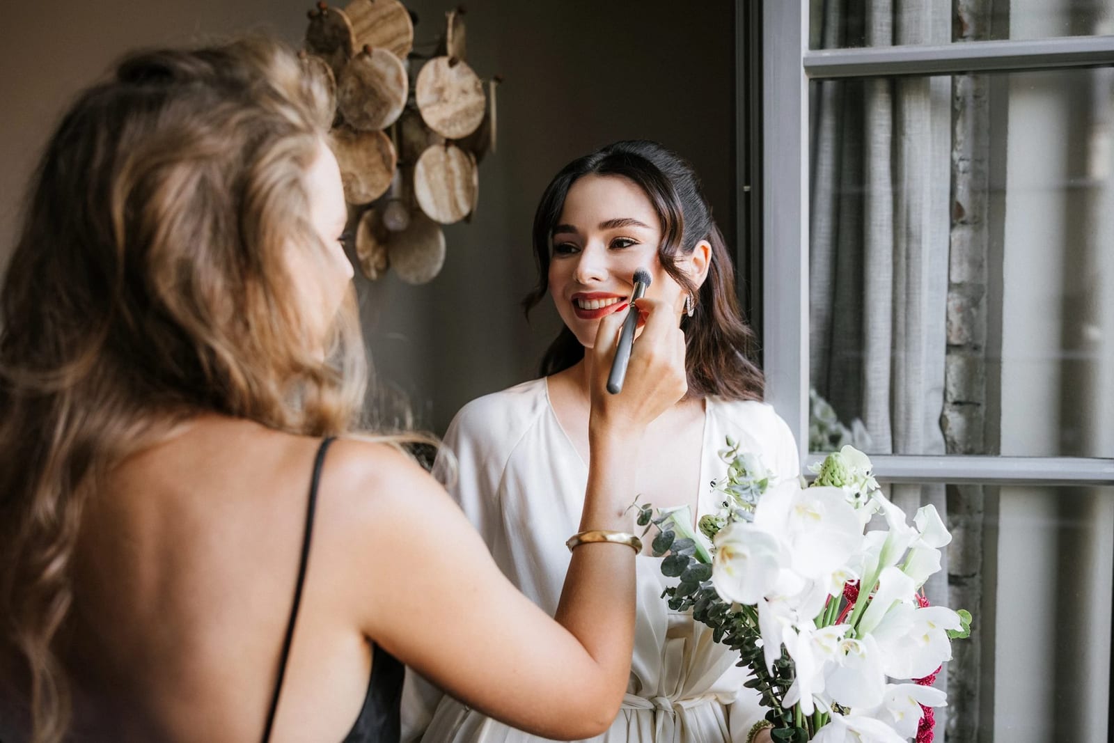 Valeria applying bridal makeup to a bride on the morning of her wedding, bouquet in hand