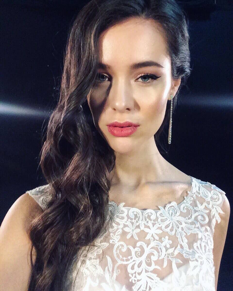 A woman with long wavy brown hair, styled by a makeup artist London expert, wears a white lace dress and a long earring, posing in front of a dark background.