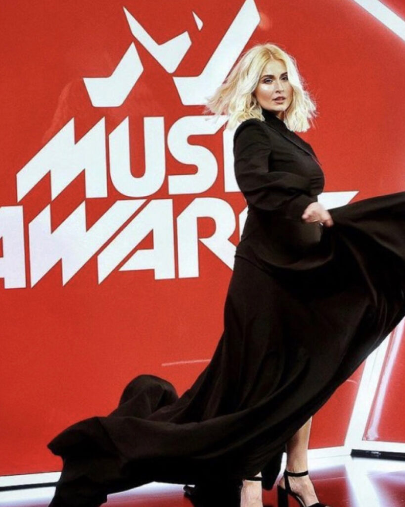 A woman in a long black dress, styled by a top makeup artist London, poses in front of a red and white "Music Awards" backdrop.