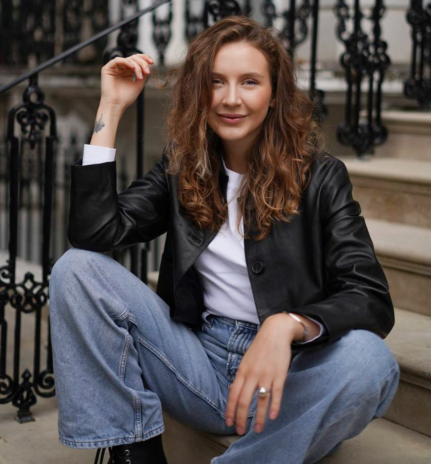 A woman with wavy brown hair sits on outdoor steps, wearing a black leather jacket, white shirt, blue jeans, and black boots, smiling at the camera. As a makeup artist London-based, her flawless look stands out against the wrought iron railings in the background.