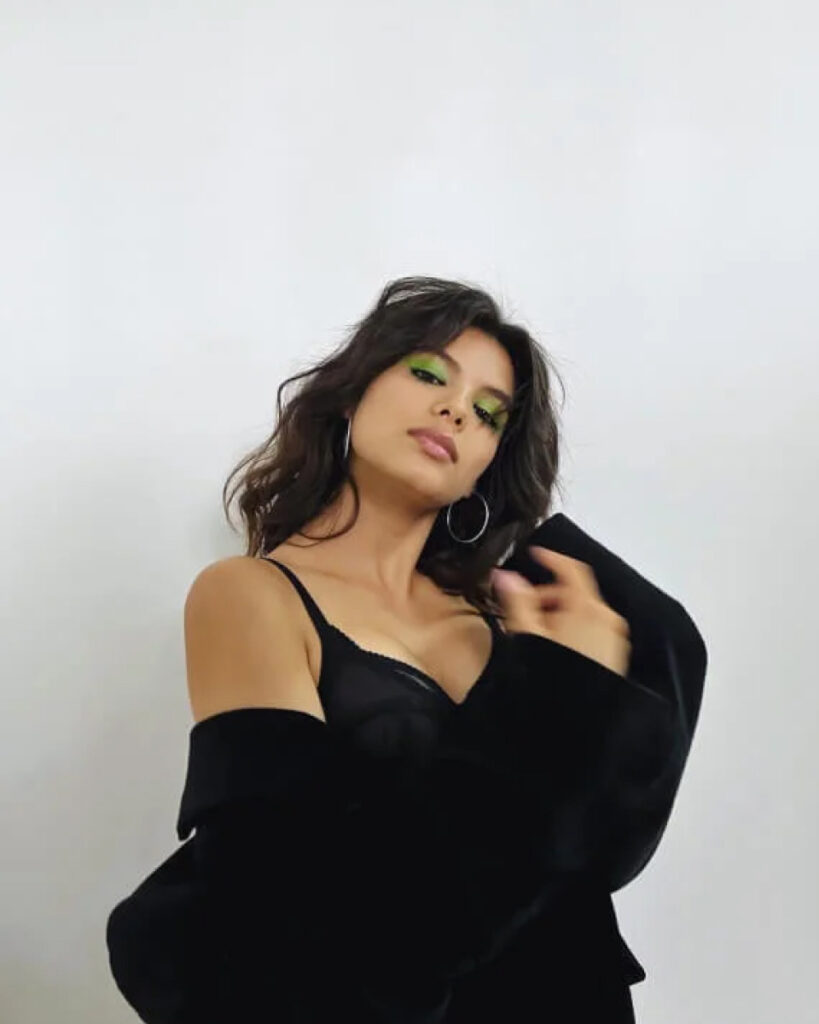 Woman with wavy brown hair wearing a black camisole and jacket poses against a plain white background, styled by a makeup artist London, featuring bold green eye makeup and hoop earrings.