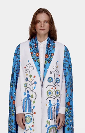 A person with long brown hair wears a white robe with colorful floral and human figure embroidery, and a blue patterned shawl draped over their shoulders.