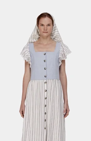 A woman stands facing forward, wearing a light blue and white striped dress with buttons down the front and a matching striped headscarf.
