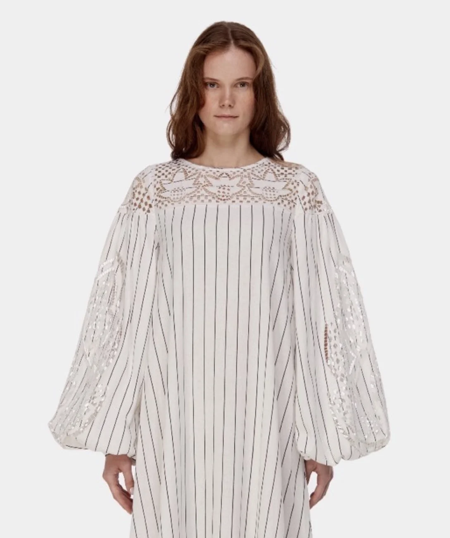 A woman wearing a white dress with vertical black stripes, lace detailing on the upper chest and sleeves, and voluminous balloon sleeves, standing against a plain background.